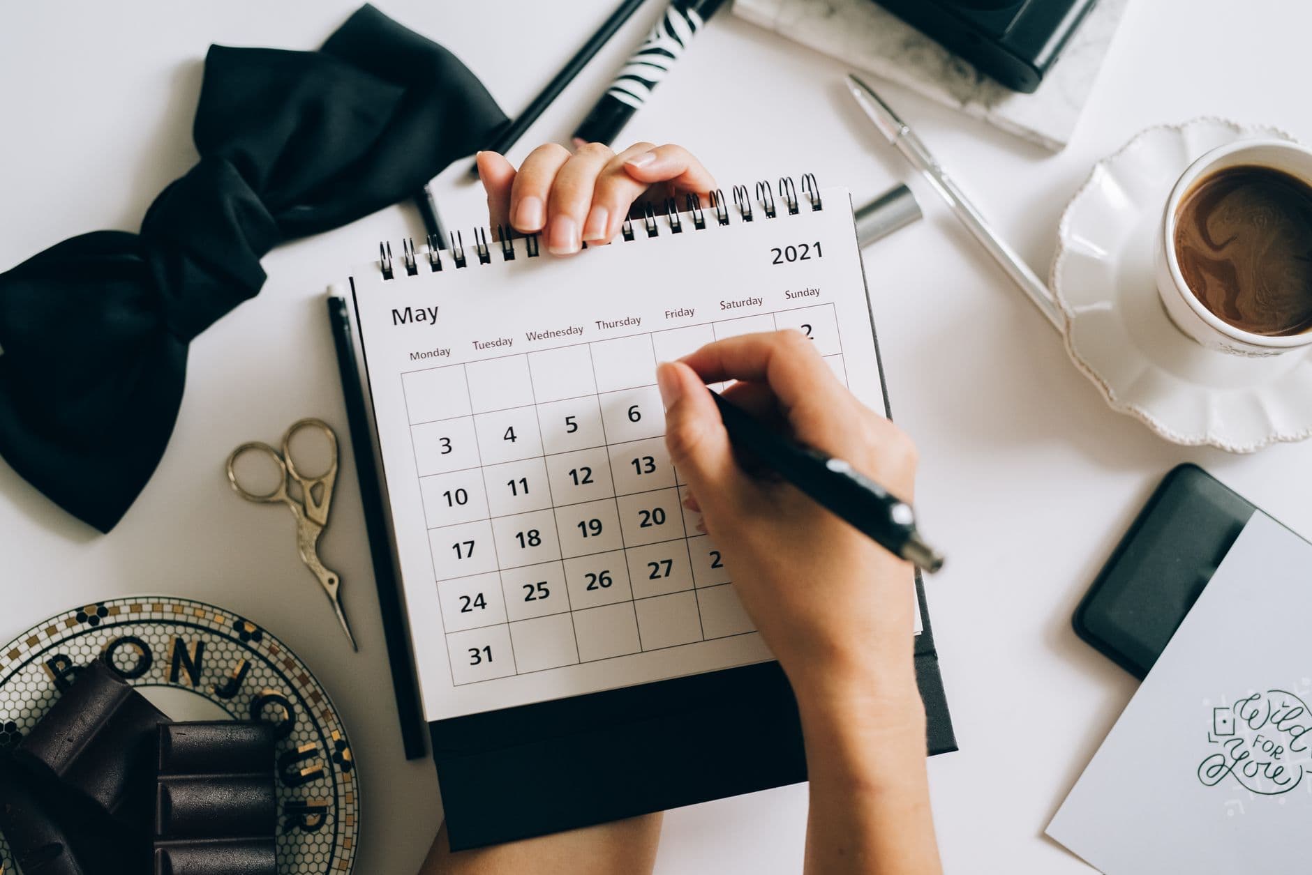 A hand marking a date on a spiral desk calendar beside a cup of coffee